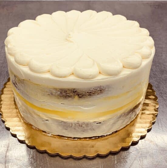 Lemon Cream Cake: pale cake with decorative frosting on the top and semi naked frosting on the sides, with a stripe of lemon curd. Cake is on a gold cake board, on a metal counter top.