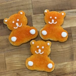 Three cute, orange sanding sugar Teddy Bear Cookies, with eyes, mouth, ears, and paws detailed in white icing, arranged on a wooden countertop background.