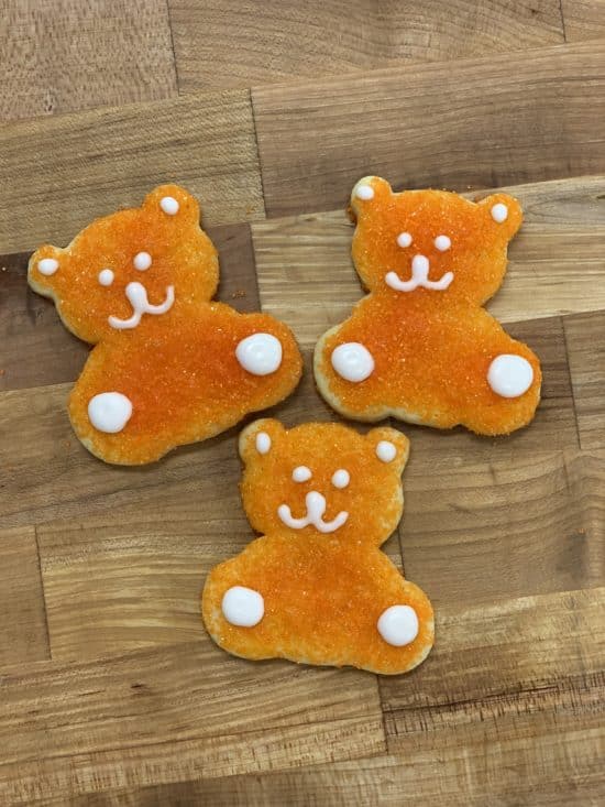Three cute, orange sanding sugar Teddy Bear Cookies, with eyes, mouth, ears, and paws detailed in white icing, arranged on a wooden countertop background.