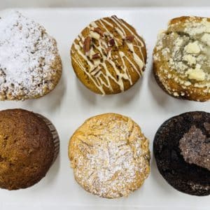 The tops of 6 muffins, in two rows of three. Top left is Cinnamon Crumb, Pumpkin Pecan, Lemon Blueberry. The next row from the left, Morning Glory, Almond Combo, and Double Chocolate.