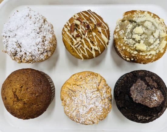 The tops of 6 muffins, in two rows of three. Top left is Cinnamon Crumb, Pumpkin Pecan, Lemon Blueberry. The next row from the left, Morning Glory, Almond Combo, and Double Chocolate.