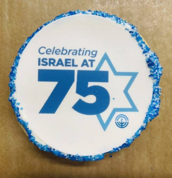 Round cookie on a brown parchment paper background. Cookie has a white fondant layer printed with a design in different blues with the text "Celebrating Israel at 75" over the outline of a Star of David and a small Jewish Federation logo at the bottom right of the star. Fondant layer has a decorative border of blue sanding sugar.