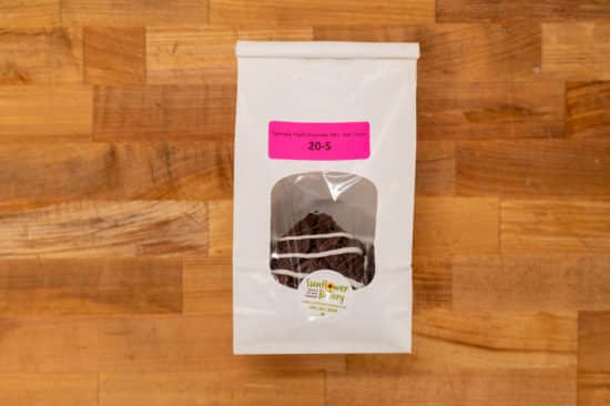 Window bag labeled "Specialty Triple Chocolate (NF) - Half-dozen 20-S" and chocolate dough hamantaschen filled with chocolate covered with chocolate drizzles visible through the window.