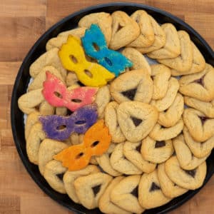 Top view of Traditional Hamantaschen Platter. 4 dozen traditional hamantaschen decoratively arrayed in the platter, garnished on the left with 5 colorful mask cookies in blue, yellow, pink, purple, and orange.