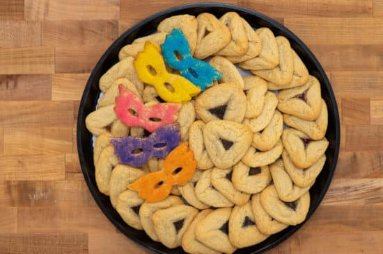 Top view of Traditional Hamantaschen Platter. 4 dozen traditional hamantaschen decoratively arrayed in the platter, garnished on the left with 5 colorful mask cookies in blue, yellow, pink, purple, and orange.