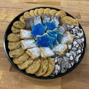 A small black platter with a ring of cookies around the rim (chocolate chip, chocolate crinkle, and snickerdoodle) and inside the ring are lemon bars, brownies, and dark blue sanding sugar cookies in the shape of the Star of David. Platter is on a wood countertop.