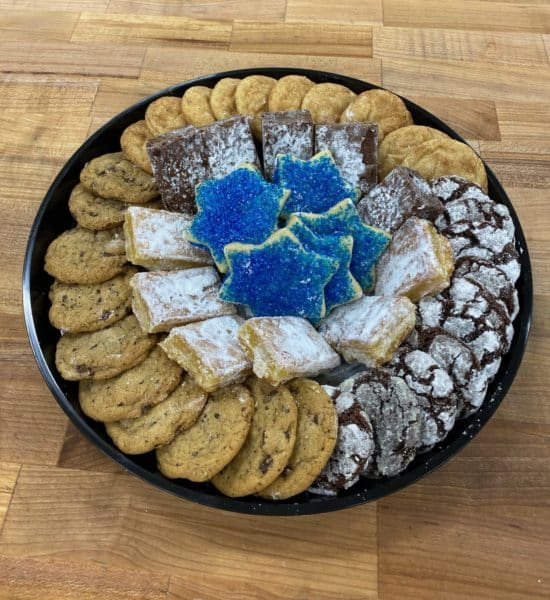 A small black platter with a ring of cookies around the rim (chocolate chip, chocolate crinkle, and snickerdoodle) and inside the ring are lemon bars, brownies, and dark blue sanding sugar cookies in the shape of the Star of David. Platter is on a wood countertop.