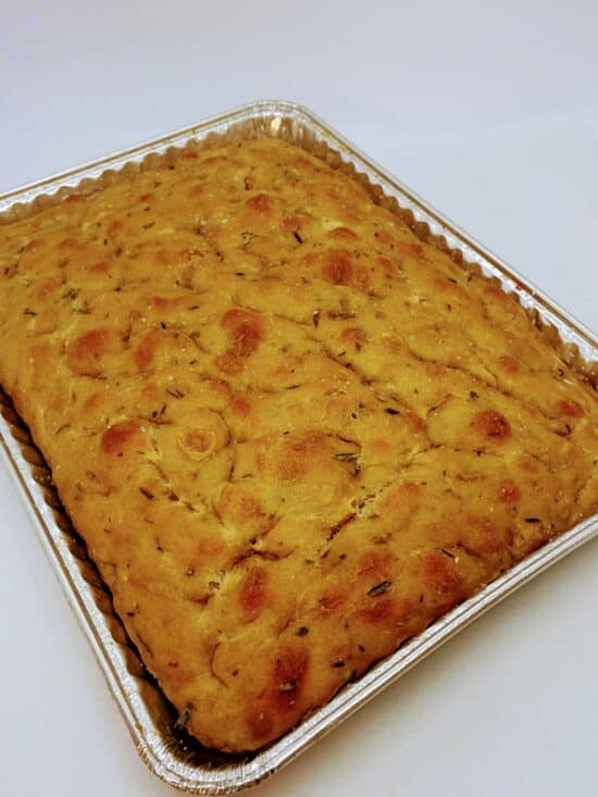 Rectangular aluminum pan with a focaccia bread with herbs mixed in and visible on the surface.