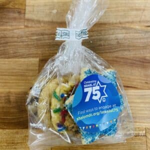 Favor bag containing one each of Star of David sanding sugar cookie, sprinkle, oatmeal raisin, chocolate chip, and snickerdoodle cookies. Bag has a sticker that reads "Celebrating Israel at 75, Find ways to engage at shalomdc.org/israelat75" Bag is on a wooden countertop.
