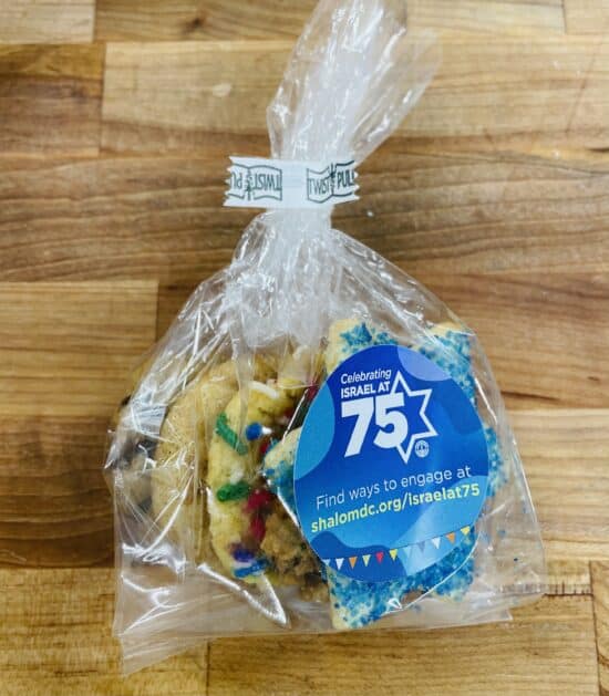 Favor bag containing one each of Star of David sanding sugar cookie, sprinkle, oatmeal raisin, chocolate chip, and snickerdoodle cookies. Bag has a sticker that reads "Celebrating Israel at 75, Find ways to engage at shalomdc.org/israelat75" Bag is on a wooden countertop.