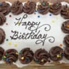 Cupcake cake with white frosting on top, with chocolate frosting dollops around the edge and colorful decorative quins. The chocolate inscription on the cake says "Happy Birthday." The cake is on a gold cake board.