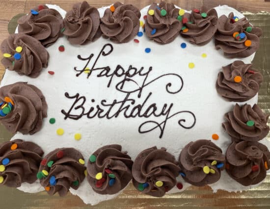 Cupcake cake with white frosting on top, with chocolate frosting dollops around the edge and colorful decorative quins. The chocolate inscription on the cake says "Happy Birthday." The cake is on a gold cake board.