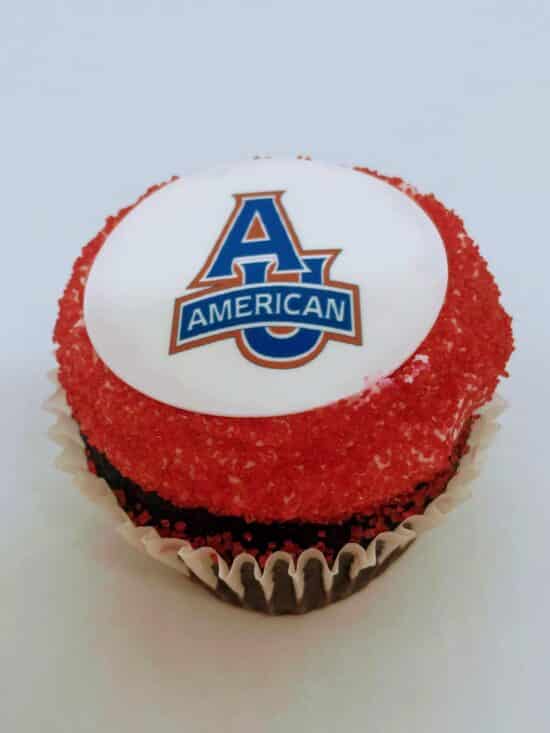 A single chocolate School Logo Cupcake, with American University Logo on a fondant disc, surrounded with red sanding sugar.