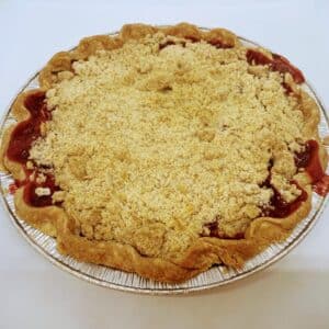 A pie with pale crumb topping, and red strawberry rhubarb filling showing at the edges. In a pie tin on a white background.