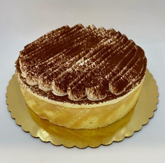 Tiramisu cake on a white background. Cake has lady finger sponge sides, has a creamy filling with a decorative ridged top, topped with cocoa powder.
