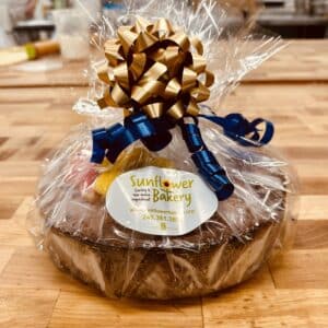 Apple Cake topped with sunflower sugar cookies, wrapped in cellophane with gold and blue ribbons, and a Sunflower Bakery sticker on the front.