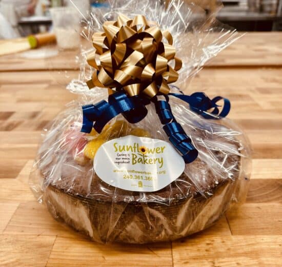 Apple Cake topped with sunflower sugar cookies, wrapped in cellophane with gold and blue ribbons, and a Sunflower Bakery sticker on the front.