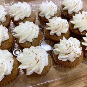 1 dozen mini cupcakes in a clear clamshell on a wooden counter. Medium brown cupcakes with a swirl of cream colored frosting on top and topped with a sprinkle of cinnamon.