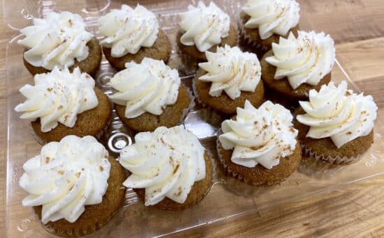 1 dozen mini cupcakes in a clear clamshell on a wooden counter. Medium brown cupcakes with a swirl of cream colored frosting on top and topped with a sprinkle of cinnamon.