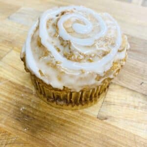 Singular muffin sitting on a wooden counter. The muffin is a light tan color, with the top coated in icing, with a decorative icing swirl.