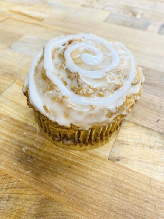 Singular muffin sitting on a wooden counter. The muffin is a light tan color, with the top coated in icing, with a decorative icing swirl.