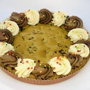 A large chocolate chip cookie cake in a paper tart form, with a decorative edge of vanilla and chocolate swirls, decorated with colorful quins.