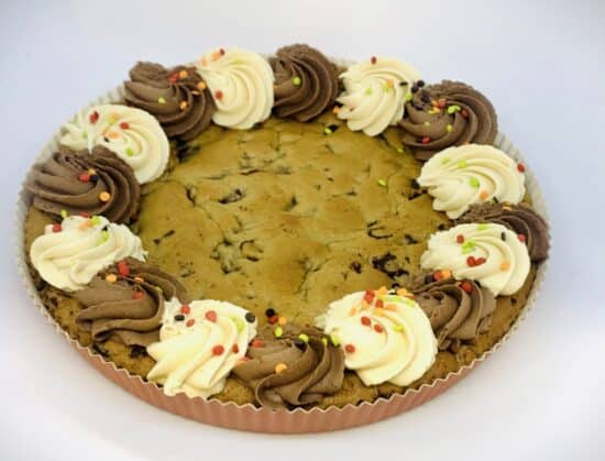 A large chocolate chip cookie cake in a paper tart form, with a decorative edge of vanilla and chocolate swirls, decorated with colorful quins.