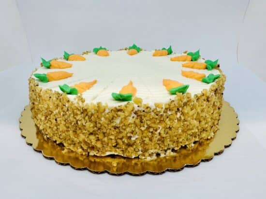 A cake with a creamy frosting top and the sides covered in walnut pieces . The top is decorated with frosting carrots, pointing inward.