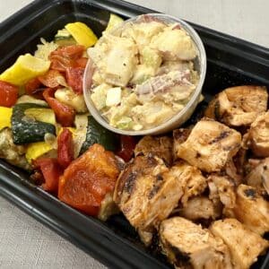To-go box with roasted veggies, chopped BBQ chicken, and a cup of potato salad.