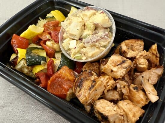 To-go box with roasted veggies, chopped BBQ chicken, and a cup of potato salad.