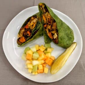 Roasted veggie wrap, diced fruit, and pickle spear on a plate.